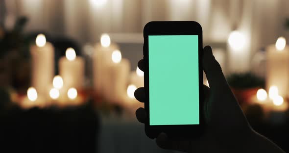 Male Hand Is Holding Smartphone with Green Screen. Lighted Candles in the Background