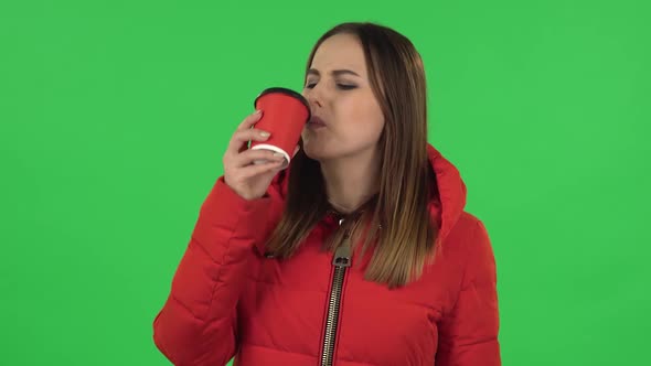 Portrait of Lovely Girl in a Red Down Jacket Is Drinking Unpalatable Coffee and Is Disgusted. Green