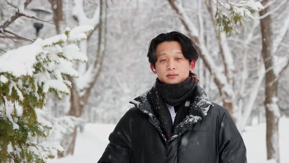 Young Asian Man in Winter Jacket Stands in Snowy Forest and Fixes His Hair