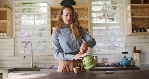 Happy caucasian woman standing at counter in cottage kitchen pouring tea from teapot and smiling