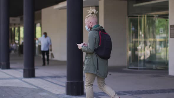 Albino african american man with face mask and dreadlocks walking and using smartphone