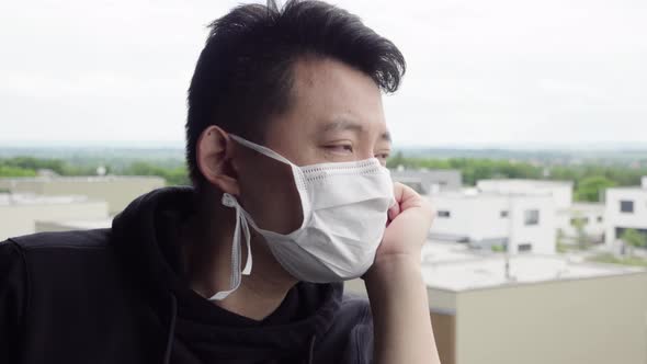 A Young Asian Man in a Face Mask Stands on a Balcony and Looks at a Cityscape Below