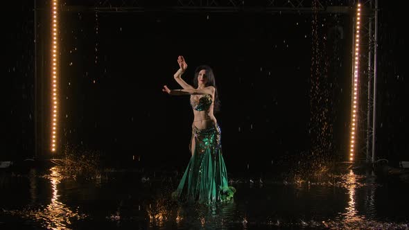 Attractive Belly Dancer Shakes Hips in Shining Oriental Costume Amid Raindrops on Surface of the