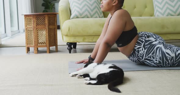 Happy african american plus size woman practicing yoga, stretching in living room with her pet cat