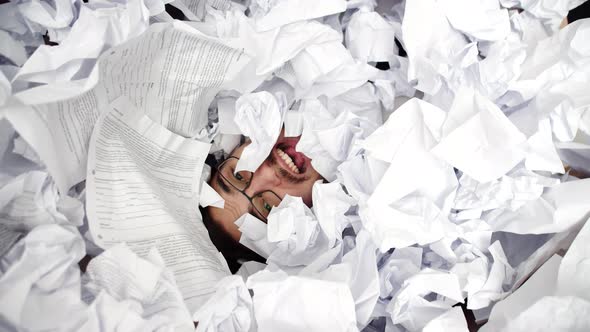 Face of male office worker screaming buried under pile of crumpled papers, top view. Man screams