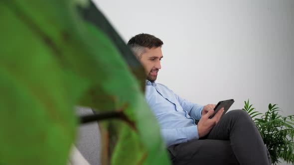 Smiling Businessman in a Light Blue Shirt Sitting on the Couch Using Tablet