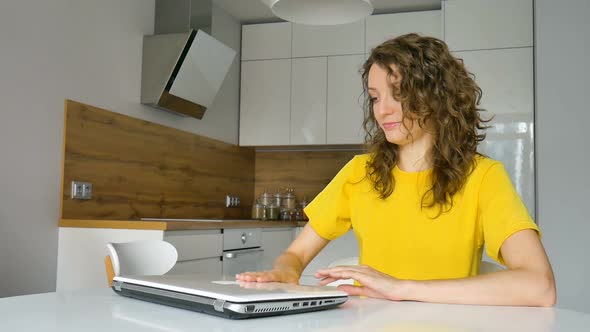 Young Woman with Curly Hair and Yellow Shirt is Working From Home Using Her Laptop at the Kitchen