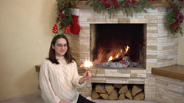 A Young Pregnant Woman in Glasses Sits By the Fireplace with a Sparkler in Her Hand