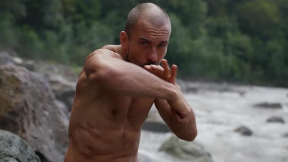an Unshaven Man with a Bare Torso is Boxing By a Mountain River