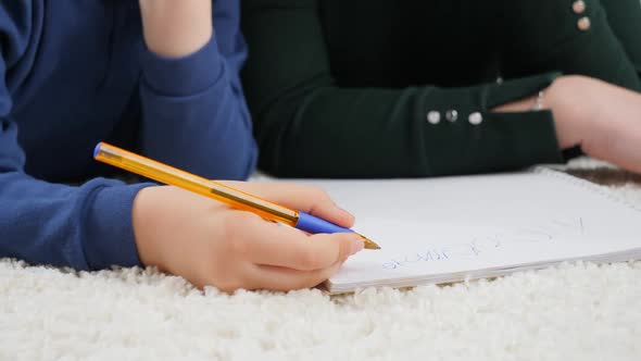 Closeup of Little Boy Writing with Pen in Copybook While Lying on Carpet in Living Room