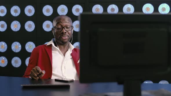 Handsome Black Guy in Red Suit Sits at Desk Listens to Music Via Headphones