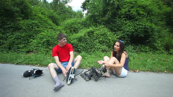 Young Woman And Man Sitting On Track, Putting Their Rollerblades On Their Feet. 1