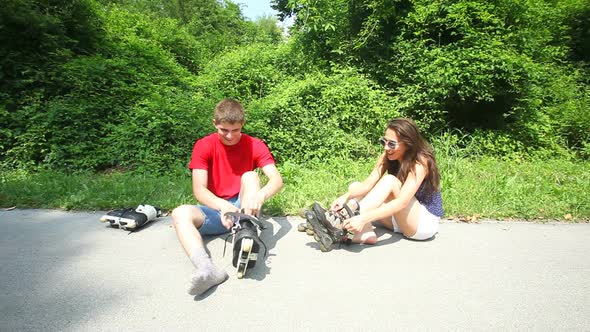 Young Woman And Man Sitting On Track, Putting Their Rollerblades On Their Feet. 2