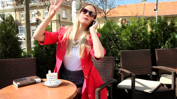 Beautiful Blond Woman Sitting In Coffee Shop, Answering The Phone And Waving alt