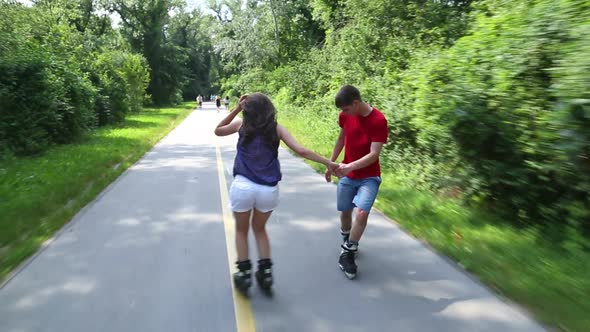 Happy Young Couple Rollerblading On A Wonderful Sunny Day In Park, Holding Hands