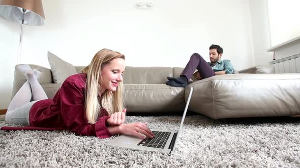 Woman Laying On Carpet Looking At Laptop With Man Sitting On Couch In Background 1