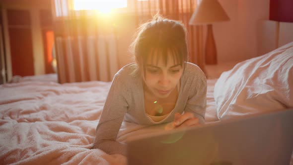 Young Woman in Pajamas Using Laptop Computer While Relaxing in Bed at Home, Sunlight in Bedroom.