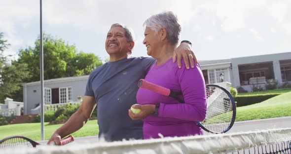 Video of happy biracial senior couple embracing and walking with rackets on tennis court