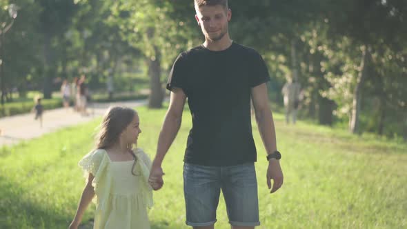 Young Caucasian Father and Little Daughter Walking in Sunshine in Spring Summer Park Holding Hands