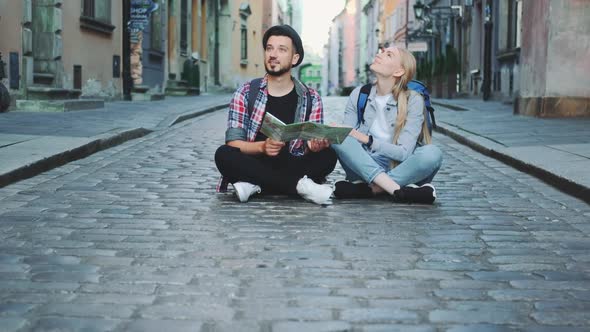 Tourists Couple with Map Sitting on Pavement and Admiring Historical Surroundings
