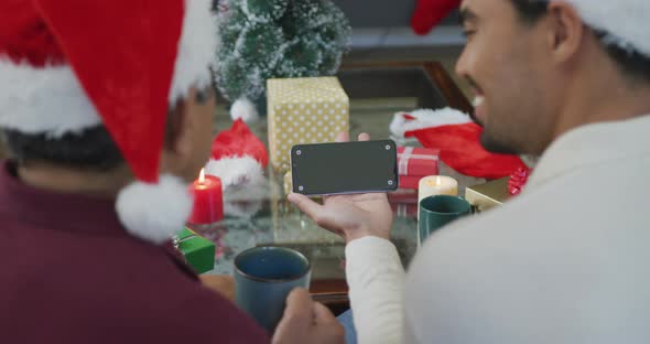 Biracial adult son and senior father in santa hats making smartphone christmas video call