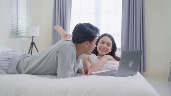 Asian young couple lying down on bed use laptop watch movie in bedroom.