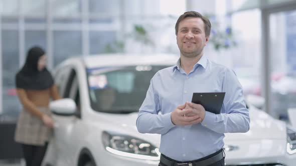 Confident Smiling Man Posing in Car Dealership with Blurred Woman in Hijab Choosing Vehicle at