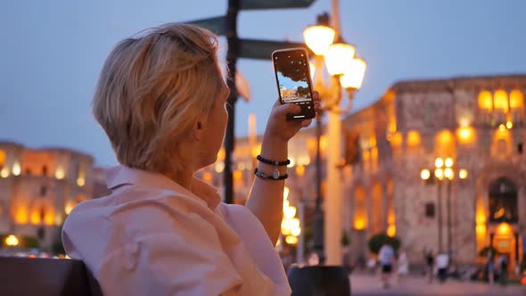 Attractive Caucasoid Blonde Sitting on a Bench Takes Pictures on a Smartphone of Beautiful City