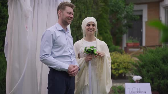 Happy Young Bride and Groom Bragging Wedding Rings on Fingers Looking Away at Altar Outdoors