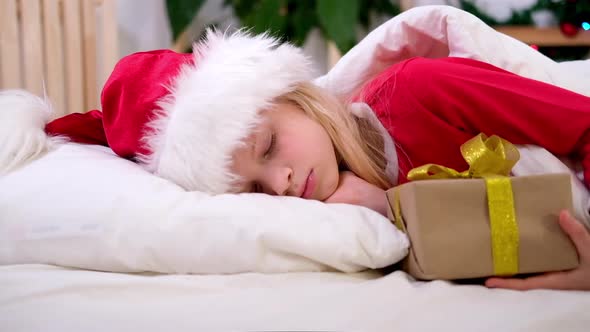 Little Girl in Santa Hat and Christmas Pajamas Lying in Bed with Gift Box at Home