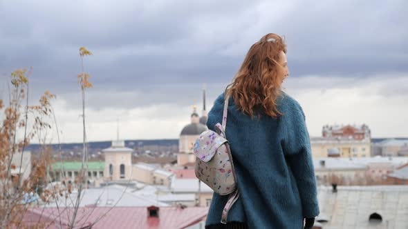 Young Redhaired Woman in a Blue Coat and Walks Against the Backdrop of the Old City