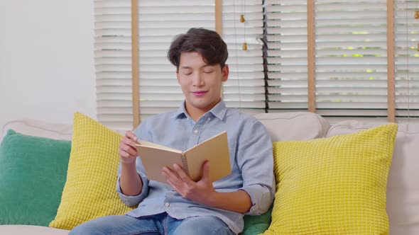 Happy handsome asian man sit on couch smile and reading book relax time at home