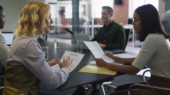 Diverse group of business colleagues wearing talking in meeting room holding documents using laptop