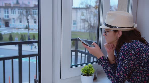 Girl in a Hat and Glasses Leaning Her Elbows on the Windowsill Looks Out the Window