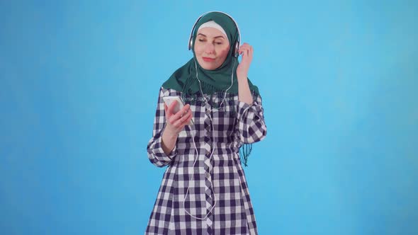 Portrait Young Muslim Woman with Headphones and a Phone Listening To Music and Dancing on a Blue