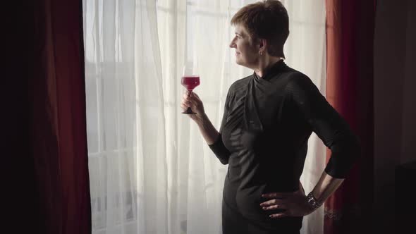 Senior Beautiful Caucasian Lady Standing in Sunrays with Glass of Wine and Looking at Window. Mature