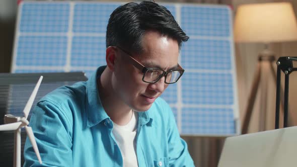 Close Up Of Asian Man Sitting In Front Of Solar Cell Looks At Wind Turbine While Typing On A Laptop