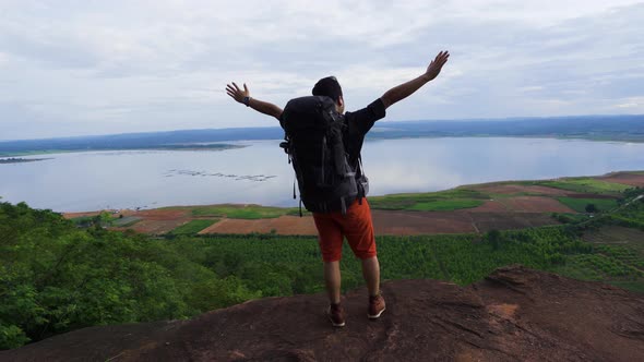 cheerful hiker man gesture raised arms on the edge of cliff, on a top of the rock mountain