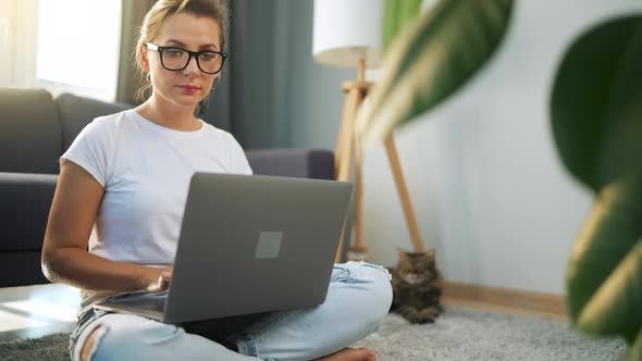 Woman with Glasses is Sitting on the Floor and Working on a Laptop