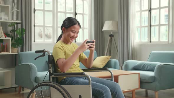 Excited Asian Kid Girl Sitting On Wheelchair Playing Video Game On Mobile Phone In House