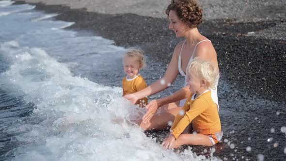 Cheerful Young Mom with Twin Blonde Toddles Sitting on the Ocean Shore Catching Waves Playing