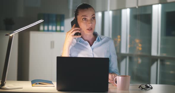 Angry Businesswoman Talking on Smartphone While Working at Laptop in Dark Office