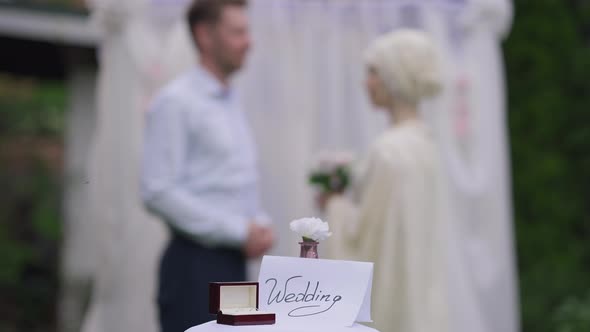 Closeup of Wedding on Paper with Rings in Box on Table and Blurred Affectionate Loving Couple at