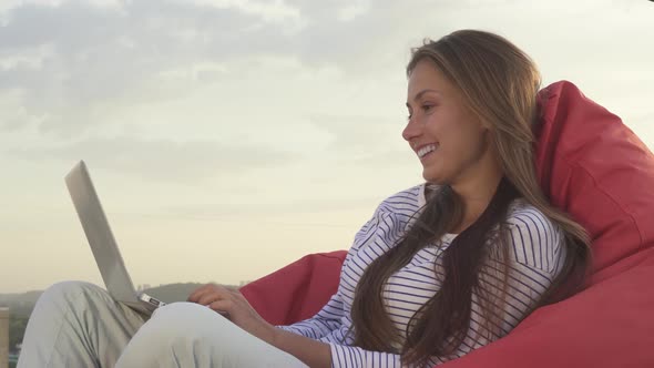 A Nice Girl Sits in a Soft Puff and Types Something on the Laptop Keyboard