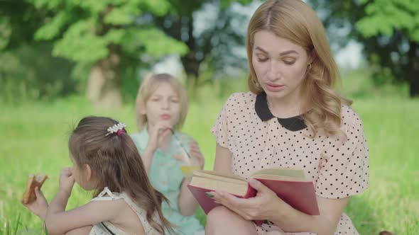 Middle Shot of Beautiful Blond Woman Reading on Sunny Meadow To Daughter and Son Sitting at the