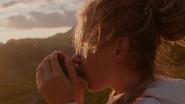 Woman Traveler with dreadlocks  drinks hot tea from thermos and looks at sunrise from mountain.