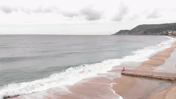 Lonely young woman sitting alone on concrete pier, looking at stormy sea with waves