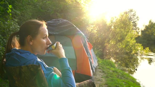 Woman is Drinking Tea in an Armchair Near a Tent on the River Bank