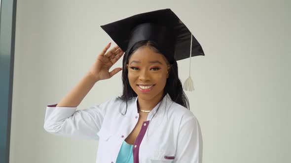 A Young African American Female Graduate in a Master's Hat and White Medical Gown Smiles Poses for