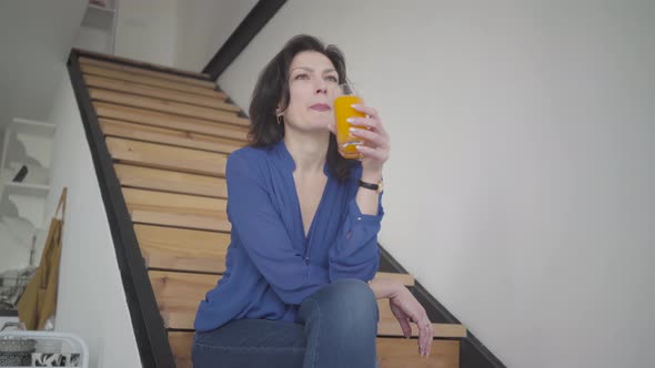 Bottom View of Elegant Caucasian Woman Drinking Orange Juice on Stairs at Home. Successful Adult